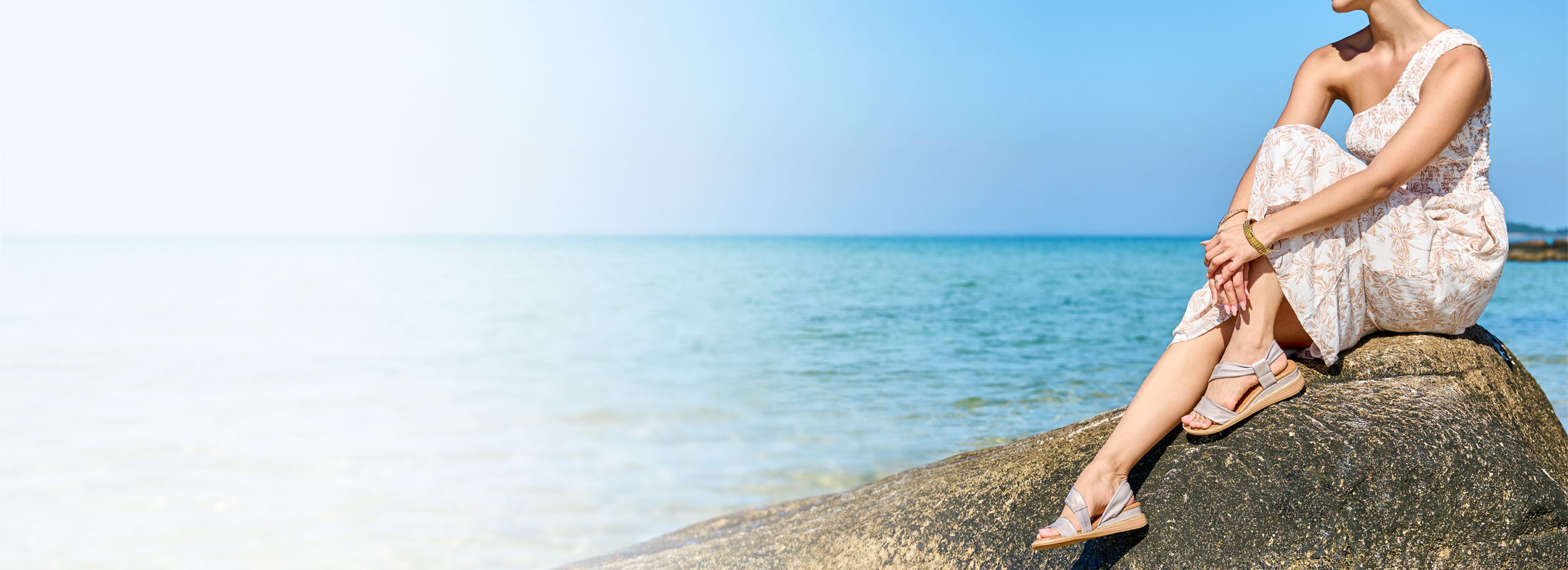 A woman sitting on a rock by the ocean wearing Marcy sandal in metallic.
